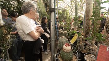 A greenhouse tour group listening to a guide while surrounded by diverse plant life, including tall cacti.