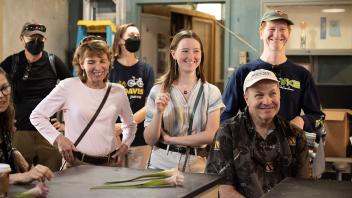A group of smiling visitors, including a young woman and her parents, engaged in a discussion at a greenhouse learning session.
