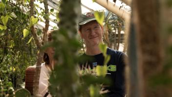A man in a green cap smiles while observing plants through the branches in a greenhouse.
