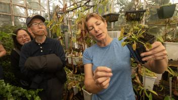 A woman in a blue shirt holds a potted Venus flytrap while explaining its features to a group of visitors in a greenhouse.