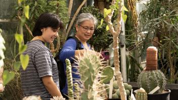 A woman and a young adult smile while looking at a smartphone, surrounded by cacti and succulents in a greenhouse.
