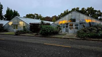 Exterior of a greenhouse at dusk with glowing orange and yellow lights visible through the translucent panels.