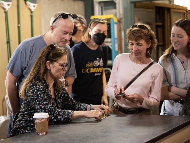 A group of people gathered around a table in a classroom setting, examining a plant specimen. A woman in a floral blouse is touching the plant, while another woman in a pink shirt holds a phone. Others in the group, including a person wearing a UC Davis shirt and a face mask, observe with interest.