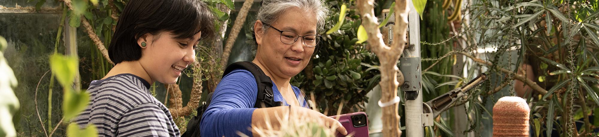 Two people smiling and observing plants inside a greenhouse, with one pointing at a plant while holding a smartphone. The background is filled with lush greenery and various plant species.