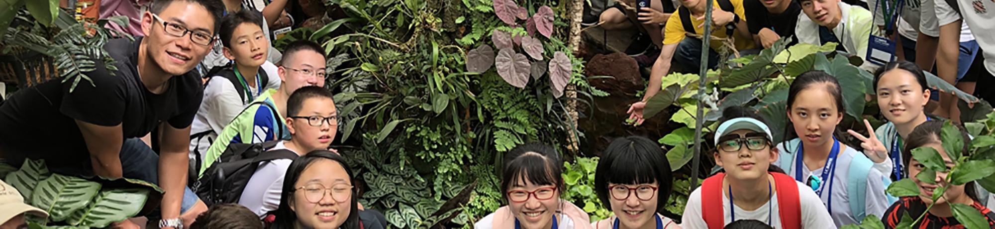 A group of students and their instructor pose together in a lush, tropical greenhouse, surrounded by vibrant green foliage and diverse plant species.